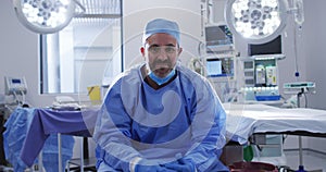 Portrait of caucasian male surgeon wearing lowered face mask sitting in operating theatre smiling