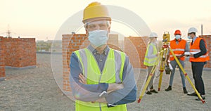 Portrait of Caucasian man constructor in casque, goggles and medical mask standing outdoors at construction and looking