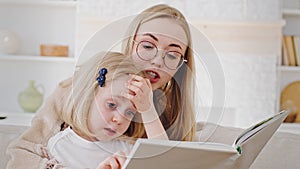 Portrait caucasian mother in glasses reads book fairy tale to child little daughter teaches baby at home, close-up