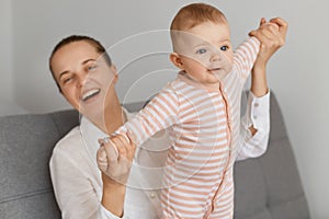 Portrait of Caucasian mother playing with her infant baby, family spending time together, woman with hair bun laughing happily