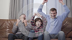 Portrait of Caucasian multi-generation family of football fans watching match on TV. Middle aged man, his adult son and