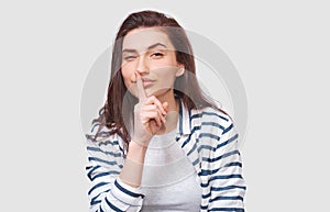 Portrait of Caucasian pretty young woman wears striped shirt, holding index finger on lips, asking to keep silence. Beautiful