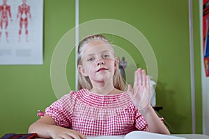 Portrait of caucasian schoolgirl sitting at desk in classroom raising hand