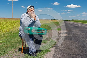 Caucasian senior having short rest on a roadside sitting on a whicker stool with an ancient green suitcase