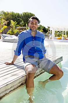 Portrait of caucasian smiling young man sitting by swimming pool at tourist resort, copy space