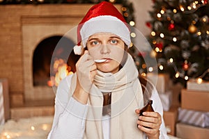 Portrait of Caucasian unhealthy woman wearing santa claus hat, being wrapped in warm scarf, sitting near Christmas tree. drinking