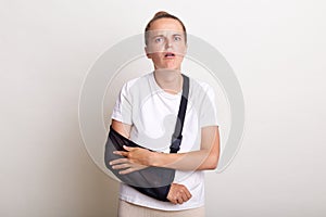 Portrait of Caucasian woman with broken arm, suffers from pain and fracture, wearing arm sling has bad mood, dressed in white t