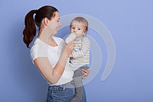 Portrait of Caucasian woman holding her baby while feeding from bottle, posing isolated ove rblue background, lady looking at her