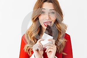 Portrait of caucasian woman 20s wearing Santa Claus red costume smiling and eating chocolate bar, isolated over white background