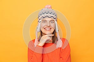 Portrait of Caucasian young adult woman wearing orange jumper and knitted earflap hat standing isolated over yellow background,