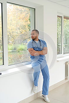 Portrait of caucasian young male doctor in blue uniform stant on hospital corridor