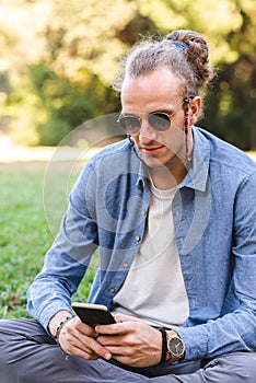 Portrait of a caucasian young man dressing causal and protecting from sun with sunglasses sitting on the grass of a green urban