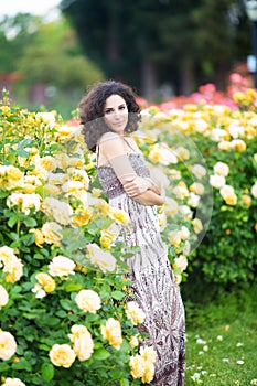 A portrait of Caucasian young woman near yellow roses bush in a rose garden, looking straight to the camera
