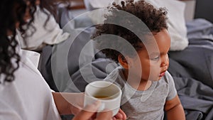 Portrait of charming African American toddler drinking healthful juice as woman feeding child. Cute handsome boy