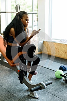 Portrait of charming black young woman with luxury long hair texting on her smartphone in the gym