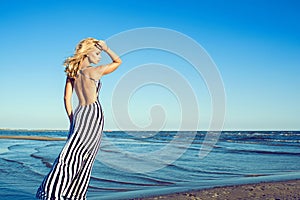 Portrait of charming blond long-haired woman in long black and white striped dress with naked back walking along the seaside