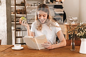 Portrait of charming blond woman reading book and eating green apple while sitting in cozy cafe indoor