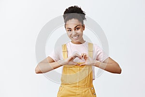 Portrait of charming and caring female dark-skinned volunteer, showing heart gesture and smiling with affection and