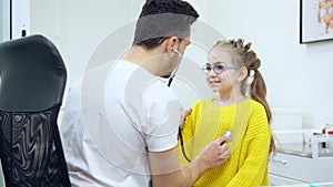 Portrait of charming Caucasian girl inhaling and exhaling as doctor using stethoscope. Cute little patient visiting