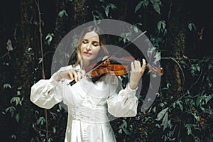 Portrait of charming Caucasian woman playing violin in tropical forest. Closed eyes. Music, art concept. Female wearing white
