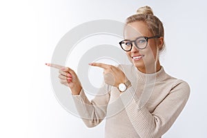 Portrait of charming cute blond european woman in glasses and sweater wearing watch pointing left and smiling inviting