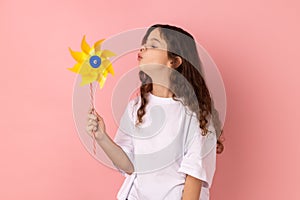 Charming playful little girl blowing at paper windmill, playing with pinwheel toy on stick.