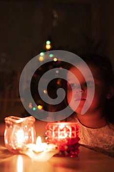 portrait of charming, dreamy girl in dark room with light from candles, Christmas lights on Christmas tree, cozy family