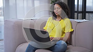 Portrait of charming happy African American young woman sitting on couch in living room typing on laptop keyboard