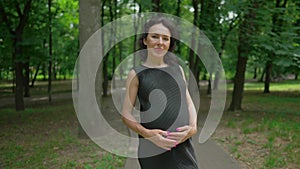 Portrait of charming happy pregnant woman standing on alley in summer spring park looking at camera smiling in slow