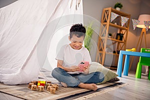 Portrait of charming happy small boy sitting floor hold gadget device wear white shirt stylish child room interior