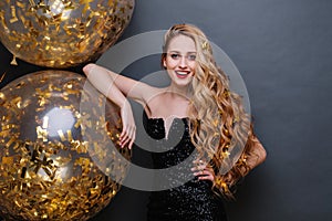 Portrait charming joyful young woman in black luxury dress, with long curly blonde hair, big balloons full with golden