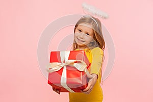 Portrait of charming kind little ginger girl with freckles and angelic halo holding big gift box, congratulating on holiday photo