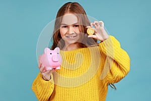 Portrait of charming little girl holds piggy bank and gold bitcoin, looking at camera with positive.
