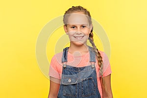 Portrait of charming little girl with long braid in denim overalls looking at camera with toothy smile