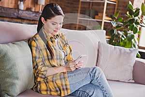 Portrait of charming peaceful girl sitting on sofa use telephone write typing social network comment house indoors
