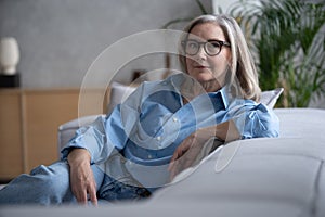 Portrait of charming senior woman with grey hair, smiling mature female sits on the couch and looks at the camera