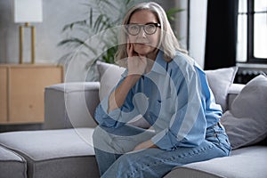 Portrait of charming senior woman with grey hair, smiling mature female sits on the couch and looks at the camera