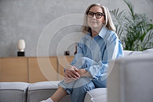 Portrait of charming senior woman with grey hair, smiling mature female sits on the couch and looks at the camera