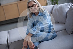 Portrait of charming senior woman with grey hair, smiling mature female sits on the couch and looks at the camera