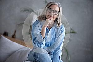 Portrait of charming senior woman with grey hair, smiling mature female sits on the couch and looks at the camera