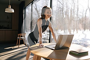 Portrait of charming sporty female in sportswear using laptop standing at desk by window on sunny day, smiling looking