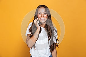 Portrait of charming woman 20s with long hair smiling and talking on mobile phone, isolated over yellow background