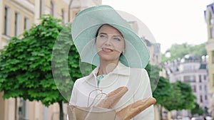 Portrait of charming woman in elegant hat standing on city street with baguettes and looking around. Beautiful Caucasian