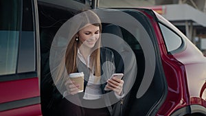 Portrait charming woman sitting inside charging electric car with modern smartphone and cup of coffee in hands. Young