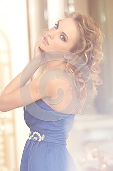 Portrait of a charming woman with stylish curly hairstyle wearing a blue dress posing next to a mirror. Beauty, vogue