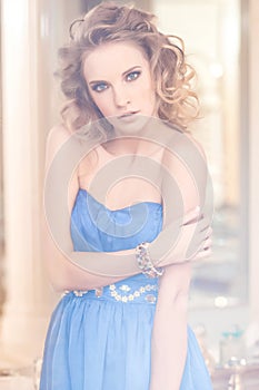 Portrait of a charming woman with stylish curly hairstyle wearing a blue dress posing next to a mirror. Beauty, vogue