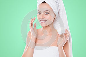 Portrait of charming woman in towel and turban on head, applying face cream for problem combined skin holding bank with