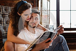 Portrait of a charming young couple at home. Woman is embracing her boyfriend near window and reading fashionable