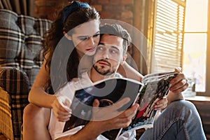 Portrait of a charming young couple at home. Woman is embracing her boyfriend near window and reading fashionable magazine on