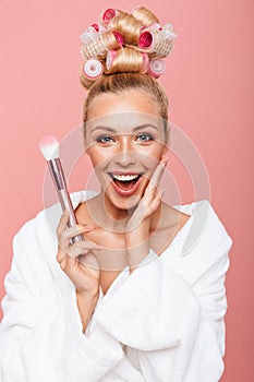 Portrait of charming young housewife in housecoat with hair curlers smiling while applying cosmetics with makeup brush photo